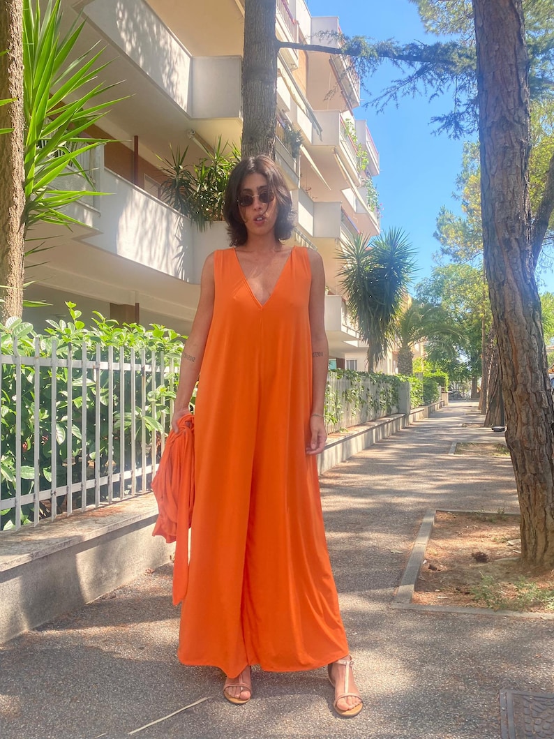 Puede incluir: Un mono naranja sin mangas con un dise&ntilde;o de pierna ancha. El mono tiene un escote en V profundo y se combina con tela naranja a juego. La persona lleva gafas de sol y sandalias. El fondo incluye &aacute;rboles, un edificio y una acera.