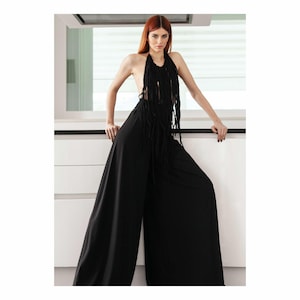 Op de afbeelding: Een vrouw draagt een zwarte haltertop met franjes en een zwarte broek met wijde pijpen. Ze staat in een moderne keuken met witte kasten.