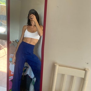 May include: A full-length mirror with a red frame reflects a person wearing a white crop top and high-waisted, wide-leg blue pants. The mirror is leaning against a white wall. A white chair is visible in the background.