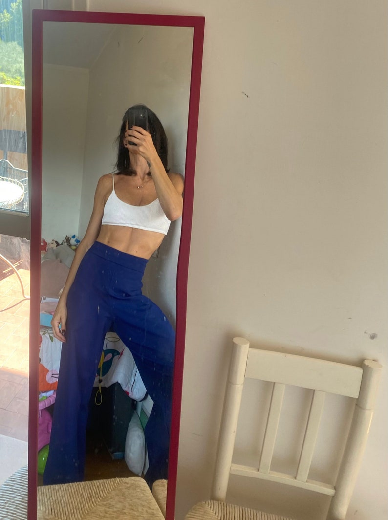 May include: A full-length mirror with a red frame reflects a person wearing a white crop top and high-waisted, wide-leg blue pants. The mirror is leaning against a white wall. A white chair is visible in the background.