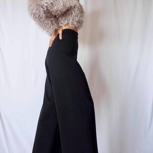 May include: A woman's outfit featuring a cropped, fluffy, light pink top and high-waisted, wide-leg black pants. The top has a textured, fringed appearance. The pants are a solid, dark color. The outfit is set against a white backdrop.