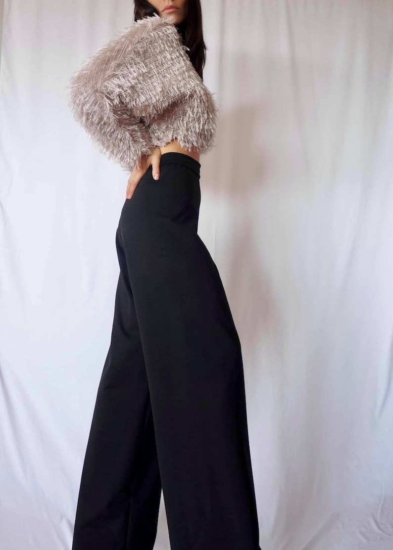 May include: A woman's outfit featuring a cropped, fluffy, light pink top and high-waisted, wide-leg black pants. The top has a textured, fringed appearance. The pants are a solid, dark color. The outfit is set against a white backdrop.