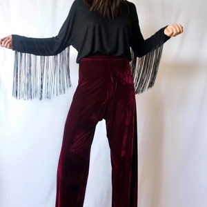 May include: A woman wearing a black long-sleeved shirt with fringe detailing on the sleeves and burgundy velvet wide-leg pants.