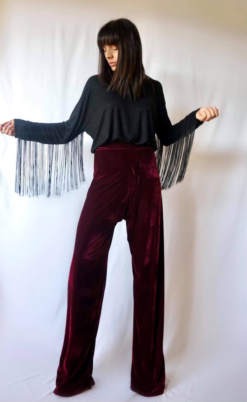 May include: A woman wearing a black long-sleeved shirt with fringe detailing on the sleeves and burgundy velvet wide-leg pants.