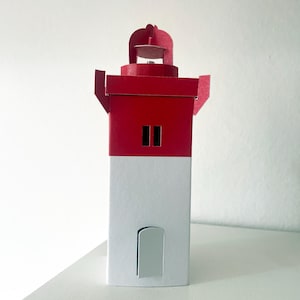 May include: A red and white paper lighthouse model. The top section is red with two black rectangular windows. The base is white with a curved door cutout. The lighthouse has a small, decorative top structure.