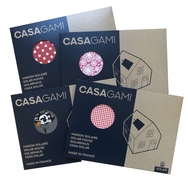 May include: Four cardboard boxes with black and white text that reads "CASAGAMI" and "MADE IN FRANCE". Each box features a different red and white patterned fabric circle. The text also reads "MAISON SOLAIRE", "SOLAR HOUSE", "SOLARHAUS", and "CASA SOLAR". The bottom right corner of the box has a small logo that reads "SOLAR".