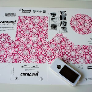 May include: A white cardboard box with instructions for building a paper lantern. The box has a pink and white floral pattern and the text "CASAGAMI la nuit maison solaire" printed on it. A small white device with a black screen is included.