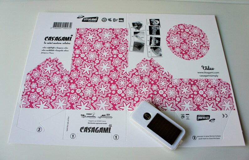May include: A white cardboard box with instructions for building a paper lantern. The box has a pink and white floral pattern and the text "CASAGAMI la nuit maison solaire" printed on it. A small white device with a black screen is included.