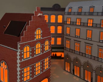 May include: Three illuminated paper model buildings with orange-lit windows. The building on the left has a red brick facade and a dark roof. The other buildings are gray and white with orange window lights, creating a warm, inviting scene.