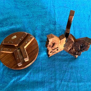 May include: A circular wooden base with three attached support legs and a separate folding wooden stand featuring intricate carved details and metal hinges. Both items are crafted from brown wood with a visible grain and natural finish.