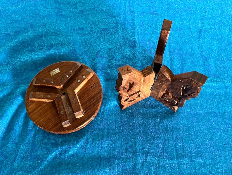 May include: A circular wooden base with three attached support legs and a separate folding wooden stand featuring intricate carved details and metal hinges. Both items are crafted from brown wood with a visible grain and natural finish.