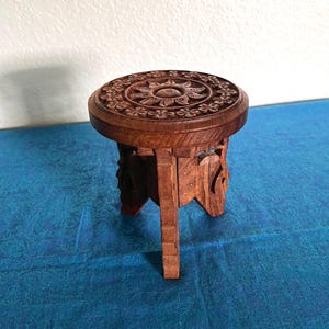 May include: A small, round, brown wooden stool featuring intricate floral and sun-like carvings on the top surface. The stool is supported by three carved legs that extend from the circular base, showcasing a natural wood grain texture throughout.