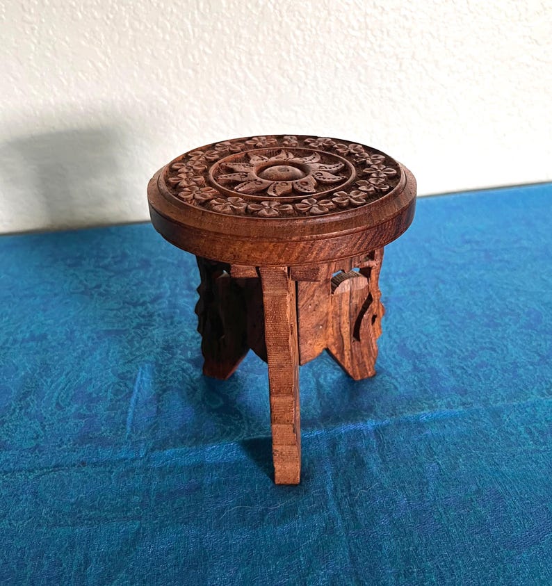 May include: A small, round, brown wooden stool featuring intricate floral and sun-like carvings on the top surface. The stool is supported by three carved legs that extend from the circular base, showcasing a natural wood grain texture throughout.