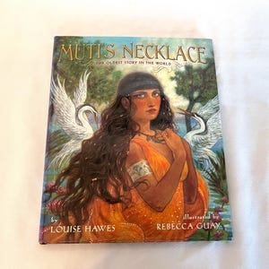 May include: A children's book titled "Muti's Necklace" with a colorful illustration of a woman in an orange dress, a necklace, and long brown hair. The cover art features two white birds and the text "The Oldest Story in the World."