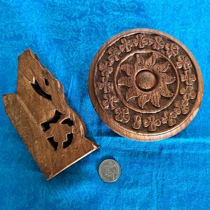May include: A brown wooden circular carved object with a floral border and sun-like centre, alongside an irregular brown wooden carved piece and a silver-coloured coin for scale. The circular piece features a repeating four-petal flower pattern.