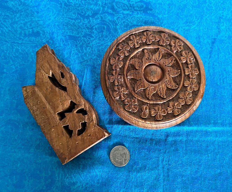 May include: A brown wooden circular carved object with a floral border and sun-like centre, alongside an irregular brown wooden carved piece and a silver-coloured coin for scale. The circular piece features a repeating four-petal flower pattern.