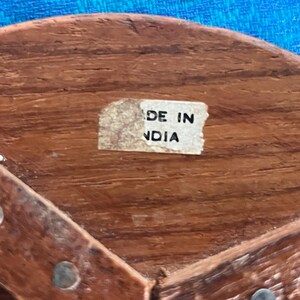 May include: A brown wooden object with a visible grain texture, featuring a small rectangular white sticker with the black text MADE IN NDIA. The surface is accented with small, evenly spaced metal studs along the curved wooden edge.