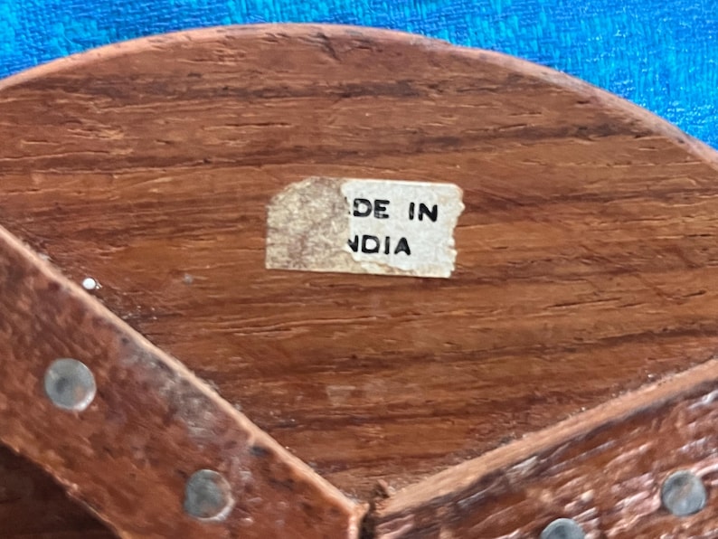 May include: A brown wooden object with a visible grain texture, featuring a small rectangular white sticker with the black text MADE IN NDIA. The surface is accented with small, evenly spaced metal studs along the curved wooden edge.