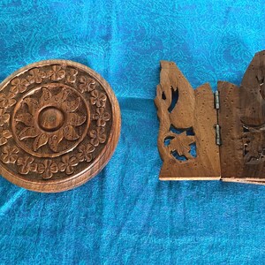 May include: A round, dark brown wooden box with a carved floral pattern on the lid, positioned next to a dark brown wooden folding stand featuring intricate cut-out leaf designs and a central metal hinge connecting the two panels.