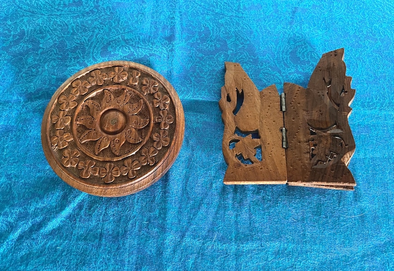 May include: A round, dark brown wooden box with a carved floral pattern on the lid, positioned next to a dark brown wooden folding stand featuring intricate cut-out leaf designs and a central metal hinge connecting the two panels.
