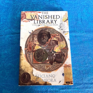 May include: A paperback book titled The Vanished Library: A Wonder of the Ancient World by Luciano Canfora. The cover features a central circular collage of ancient bronze-toned portraits and coins against a cream background with torn paper edges.