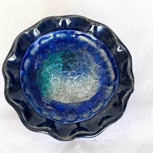 May include: A circular bowl with a ruffled edge featuring a deep navy blue outer rim and a center filled with a mosaic of translucent glass pieces in shades of cobalt blue, teal, and clear white, creating a textured and reflective surface.