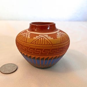 May include: A small, round, terracotta-colored ceramic pot featuring a wide opening and a short neck. The exterior is decorated with horizontal bands of geometric patterns in white, orange, and blue. A US quarter coin is placed next to the pot.