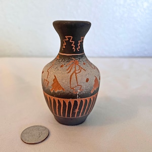 May include: A small, dark-colored ceramic vase featuring a light-colored midsection decorated with orange-toned etched figures, including a flute player, and vertical orange lines at the base. A silver-colored coin is placed next to the vase.