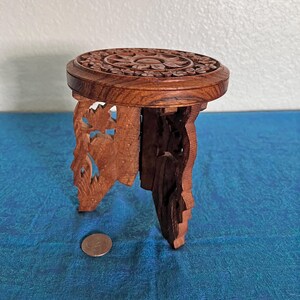 May include: A small, round, brown wooden stool featuring intricate floral carvings on the top surface and openwork leaf patterns on the three legs. A circular metal coin is placed on the surface next to the stool for scale reference.