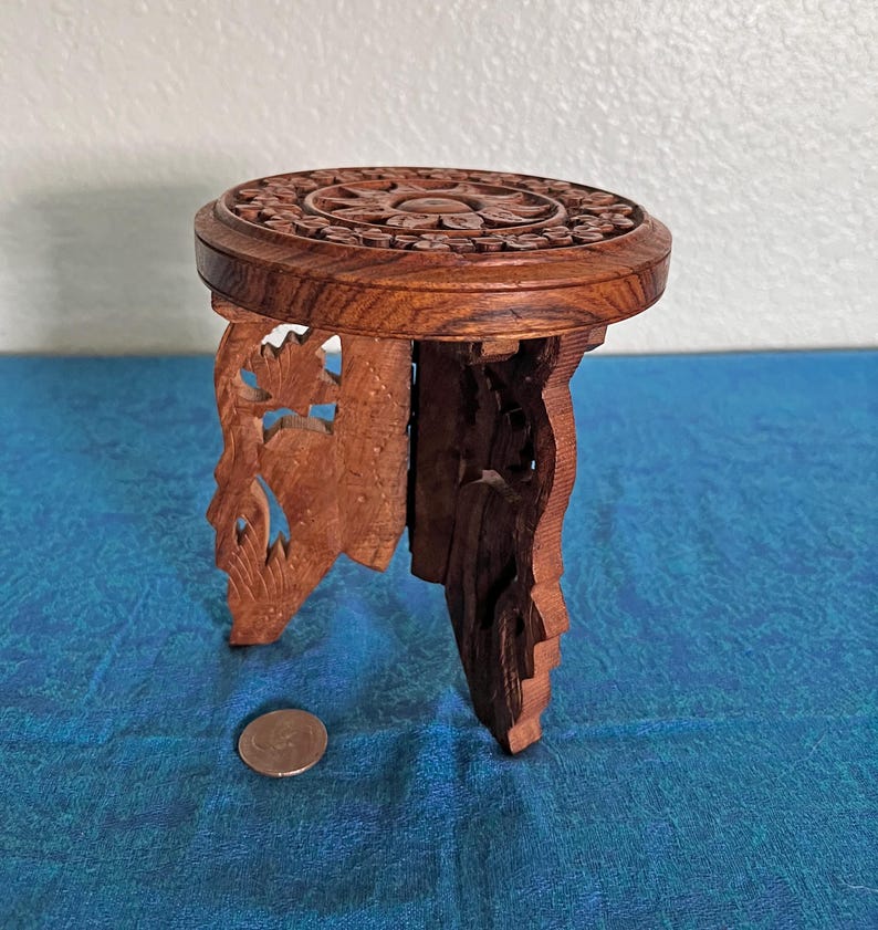 May include: A small, round, brown wooden stool featuring intricate floral carvings on the top surface and openwork leaf patterns on the three legs. A circular metal coin is placed on the surface next to the stool for scale reference.