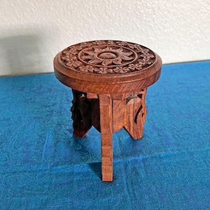 May include: A small, round, brown wooden stool featuring intricate floral and sun-like carvings on the top surface. The stool is supported by three carved legs that provide a stable base, showcasing a natural wood grain texture throughout the piece.