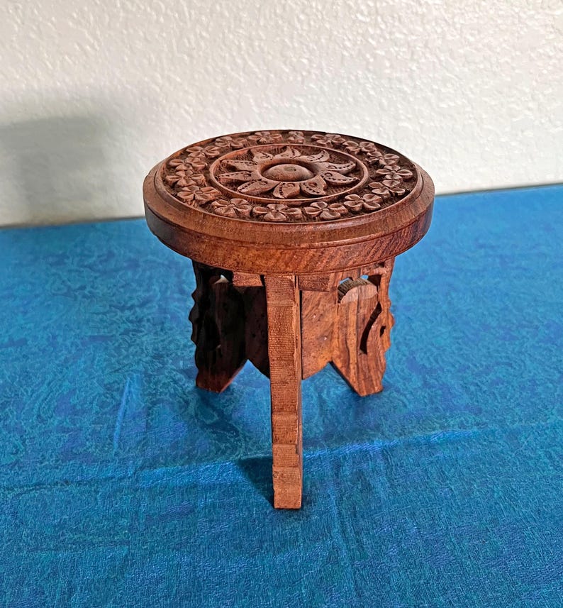 May include: A small, round, brown wooden stool featuring intricate floral and sun-like carvings on the top surface. The stool is supported by three carved legs that provide a stable base, showcasing a natural wood grain texture throughout the piece.