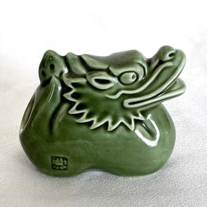 May include: A glossy sage green ceramic dragon figurine featuring a stylized head with an open mouth, prominent facial ridges, and a smooth, rounded body. A small, square black stamp with intricate markings is visible on the lower left side.