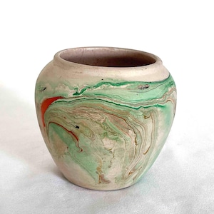 May include: A small, round ceramic vase with a marbled design. The vase features swirls of green, brown, and orange against a cream-colored background. The interior is a muted purple color. The vase has a wide opening and a slightly curved shape.