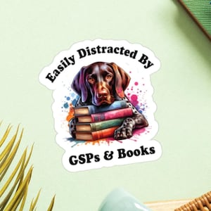 Op de afbeelding: Een bruine en witte Duitse Korthaar zit met een stapel boeken voor zich. De hond is omringd door kleurrijke verfspatten. De tekst "Easily Distracted By GSPs & Books" is in zwart op een witte achtergrond geschreven.