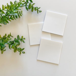 May include: Three blank white square cards on a white surface with a sprig of green foliage in the corner.