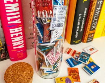 Puede incluir: Un tarro de vidrio lleno de portadas de libros en miniatura. El tarro está sentado sobre una superficie blanca con una tapa de corcho al lado. Varias portadas de libros en miniatura están esparcidas alrededor del tarro. Las portadas de los libros presentan varios títulos, que incluyen "The Henna Artist", "Unmasked", "The Cold Case", "My Life in the Lake", "Housemaid", "Kite Runner", "5 Love Languages", "Help", "The Seven Husbands of Evelyn Hugo", and "The Flames of Silver".