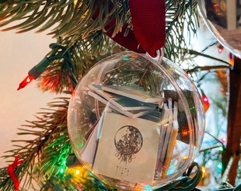 May include: A clear, spherical plastic ornament filled with miniature books, featuring a dark circular graphic on the front-most book cover. The ornament is suspended by a deep red ribbon and hangs from a green pine branch with colorful lights.