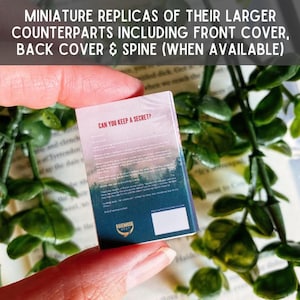 May include: A miniature book replica titled Can You Keep a Secret? featuring a forest landscape graphic on the cover, dark blue and pink text, and a small white square at the bottom. The cover includes the Ravenhood text and logo.