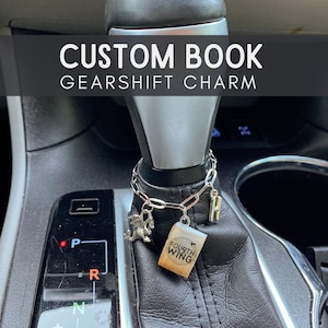 May include: A silver gearshift charm with a book, dragon, and initial pendant. The book charm reads "Fourth Wing" and "Rebecca Yarros". The charm is attached to a car's gearshift. The text "CUSTOM BOOK GEARSHIFT CHARM" is displayed above the gearshift.