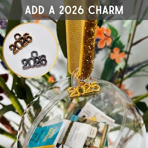 May include: A clear spherical ornament hanging from a gold ribbon, featuring a gold 2025 charm. An inset circle displays two additional metal charms with the text 2026 in gold and silver tones. The text ADD A 2026 CHARM appears at the top.