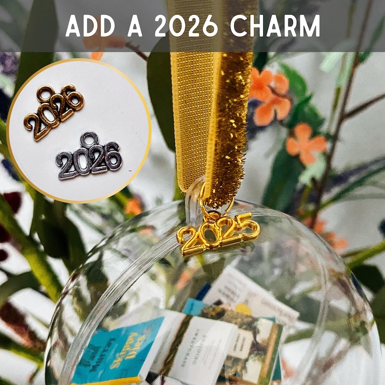 May include: A clear spherical ornament hanging from a gold ribbon, featuring a gold 2025 charm. An inset circle displays two additional metal charms with the text 2026 in gold and silver tones. The text ADD A 2026 CHARM appears at the top.
