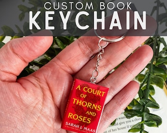 Op de afbeelding: Een miniatuur boeksleutelhanger met een rode kaft waarop de titel A Court of Thorns and Roses en de auteur Sarah J. Maas in gele tekst zijn gedrukt. De rechthoekige bedel is met een kort kettinkje bevestigd aan een zilverkleurige metalen ring.