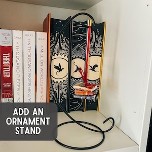 May include: A black metal ornament stand with a curved hook and a circular base. A miniature stack of three books hangs from the hook by a red ribbon. The stand is positioned in front of several books with black and gold spines featuring dragon illustrations.