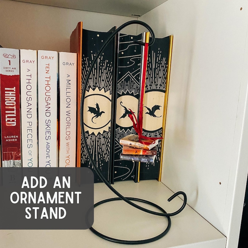 May include: A black metal ornament stand with a curved hook and a circular base. A miniature stack of three books hangs from the hook by a red ribbon. The stand is positioned in front of several books with black and gold spines featuring dragon illustrations.