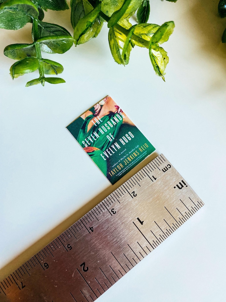Puede incluir: Un libro de bolsillo titulado "Los Siete Maridos de Evelyn Hugo" de Taylor Jenkins Reid. La portada muestra a una mujer con un vestido verde y una flor roja en el pelo.
