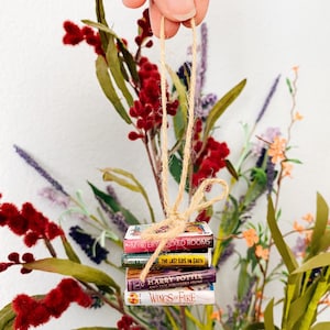 May include: A miniature stack of four books tied together with a tan twine loop for hanging. The book titles visible are Mystery Locked Rooms, The Last Kids on Earth, Harry Potter and the Sorcerer's Stone, and Wings of Fire, featuring colourful covers.
