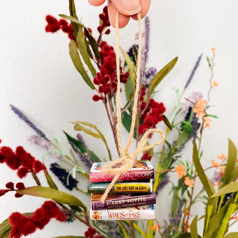May include: A miniature stack of four books tied together with a tan twine loop for hanging. The book titles visible are Mystery Locked Rooms, The Last Kids on Earth, Harry Potter and the Sorcerer's Stone, and Wings of Fire, featuring colourful covers.