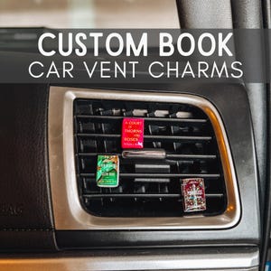 May include: Three miniature book-shaped car vent charms attached to a black plastic vehicle air vent. The charms feature covers for A Court of Thorns and Roses, The Seven Husbands of Evelyn Hugo, and Powerless. Text reads CUSTOM BOOK CAR VENT CHARMS.