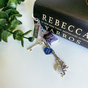 May include: A silver-toned metal keychain featuring multiple charms: a rectangular tag engraved with the word FEARLESS, a miniature book titled Onyx Storm, a silver skeleton key, a blue heart charm with the letter H, and a detailed dragon figure.